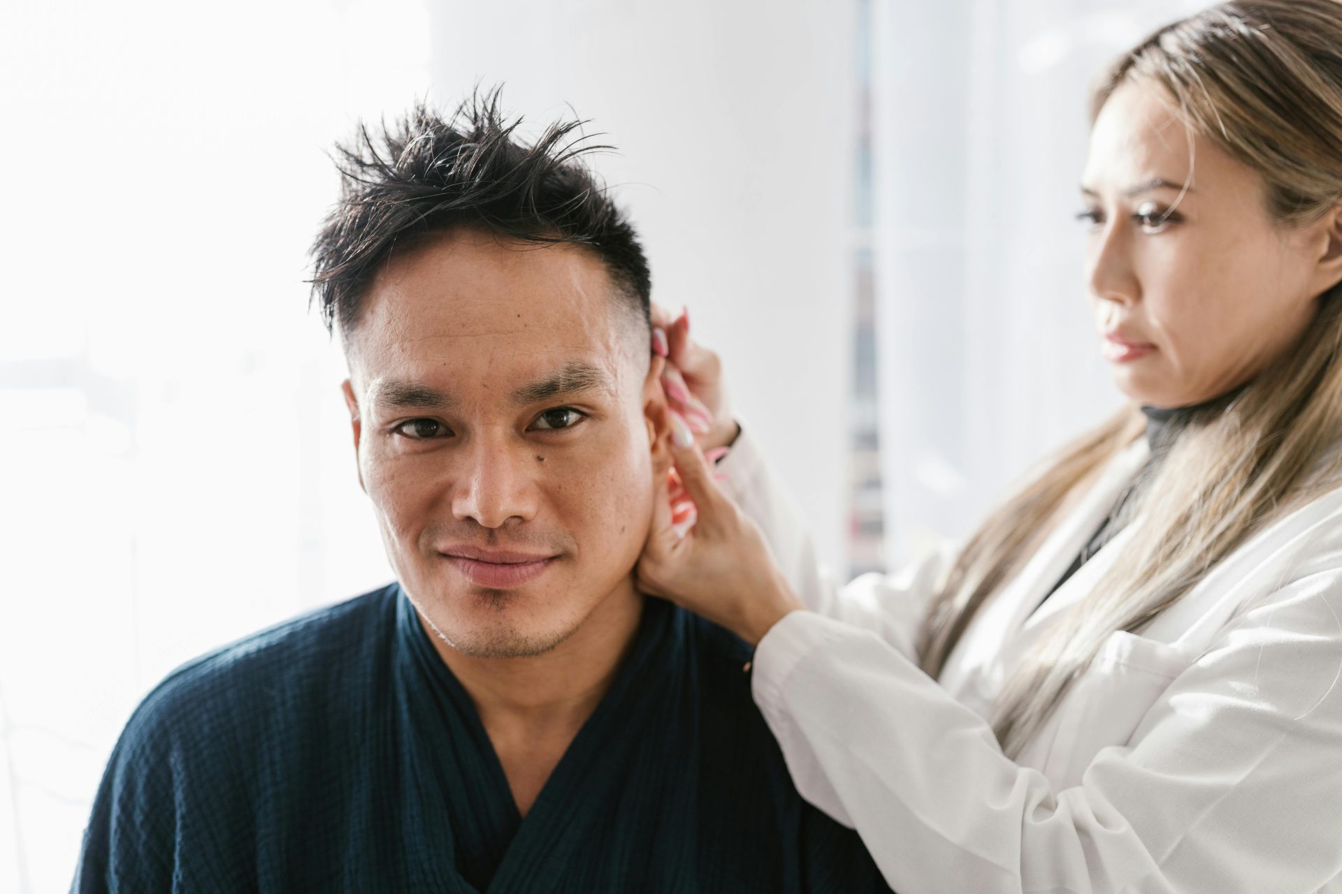 A doctor performs an ear examination on a patient in a well-lit clinic setting.